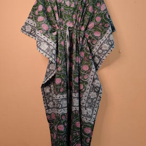 Puede incluir: Un vestido caftán largo hasta el suelo con fondo gris y un patrón floral repetitivo en tonos rosa y verde. La prenda tiene escote en V, mangas anchas estilo kimono y un cordón ajustable en la cintura con un patrón de rayas blancas y negras.