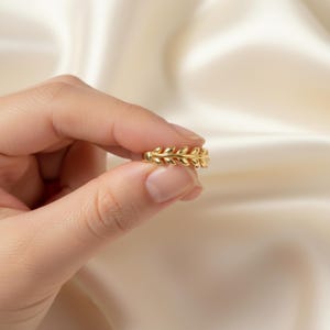 May include: A gold ring with a leaf pattern. The ring is held between fingers against a soft, cream-coloured background. The ring's design features a series of leaf-shaped elements, creating a delicate and natural aesthetic.