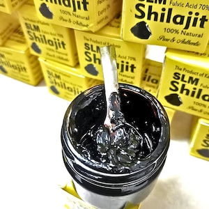Peut inclure: Un petit pot noir rempli d'une substance épaisse, sombre et visqueuse, avec une cuillère en métal argenté insérée au centre. Le pot est entouré de plusieurs boîtes jaunes étiquetées SLM Shilajit, 100% Natural, Pure & Authentic, et Fulvic Acid 70%.