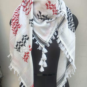May include: White scarf featuring red and black geometric patterns. The scarf has a fringed edge and white tassels. The design includes red and black zig-zag and diamond shapes. The scarf is draped over a black mannequin.
