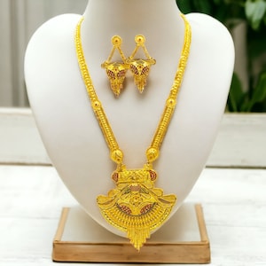Puede incluir: Un collar de oro con un gran colgante y pendientes a juego. El colgante tiene forma de abanico con detalles intrincados y piedras preciosas rojas y verdes. El collar y los pendientes están en un expositor blanco.