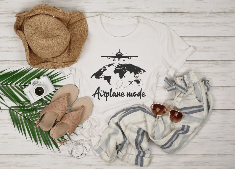 May include: A white t-shirt with a black graphic of an aeroplane above a world map and the words "Airplane mode". The image also includes a straw hat, sandals, sunglasses, a camera, and striped trousers.