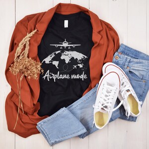 May include: A black t-shirt with the words "Airplane mode" and a graphic of a plane over a world map. The shirt is paired with blue jeans, white trainers, and a rust-coloured blazer. Dried flowers add a decorative touch.