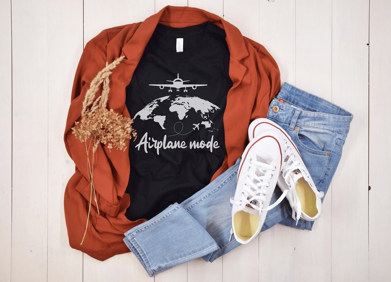 May include: A black t-shirt with the words "Airplane mode" and a graphic of a plane over a world map. The shirt is paired with blue jeans, white trainers, and a rust-coloured blazer. Dried flowers add a decorative touch.