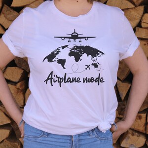 May include: White t-shirt featuring a black world map, aeroplane, and the words "Airplane mode". The shirt has short sleeves and is knotted at the waist. The background is a stack of logs.