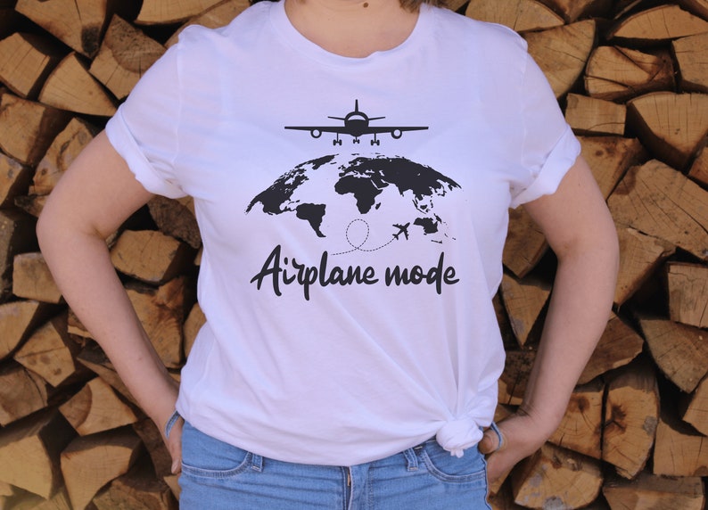 May include: White t-shirt featuring a black world map, aeroplane, and the words "Airplane mode". The shirt has short sleeves and is knotted at the waist. The background is a stack of logs.