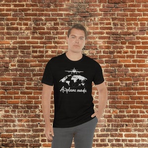 May include: Black t-shirt featuring a white graphic of an aeroplane above a world map and the words "Airplane mode". The shirt is worn by a person in front of a brick wall.