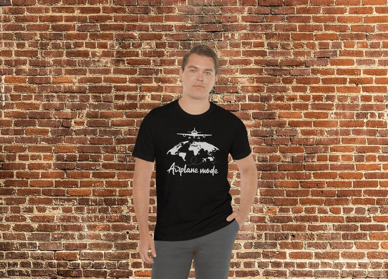 May include: Black t-shirt featuring a white graphic of an aeroplane above a world map and the words "Airplane mode". The shirt is worn by a person in front of a brick wall.