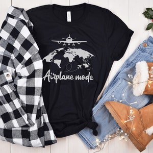 May include: Black t-shirt with a world map and airplane graphic, the words "Airplane mode" are written below. The shirt is paired with a black and white checked shirt, blue jeans, and brown boots.