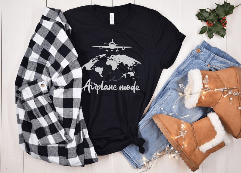May include: Black t-shirt with a world map and airplane graphic, the words "Airplane mode" are written below. The shirt is paired with a black and white checked shirt, blue jeans, and brown boots.