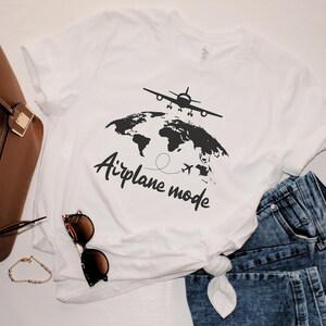May include: White t-shirt featuring a black graphic of an aeroplane above a world map, with the words "Airplane mode". The shirt is styled with blue jeans, a brown handbag, sunglasses, and a bracelet.