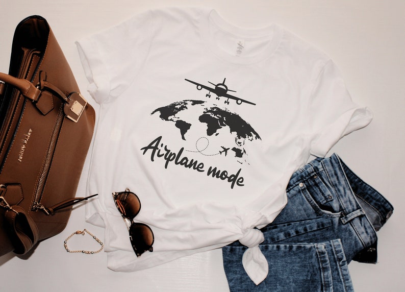 May include: White t-shirt featuring a black graphic of an aeroplane above a world map, with the words "Airplane mode". The shirt is styled with blue jeans, a brown handbag, sunglasses, and a bracelet.