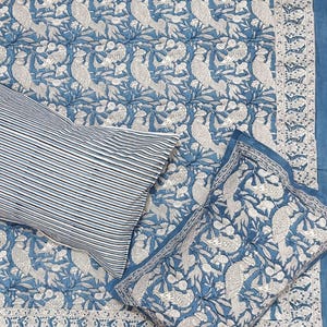 May include: A blue and white bedding set featuring a bird and floral pattern. The set includes a patterned quilt, a striped pillowcase, and a matching pillow cover. The quilt has a detailed border.