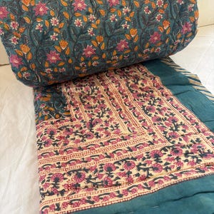 Puede incluir: Un edredón turquesa con estampado floral con flores rosas y naranjas. El edredón está doblado, revelando una sección crema con un diseño floral repetido y un borde turquesa. El edredón está hecho de material suave.