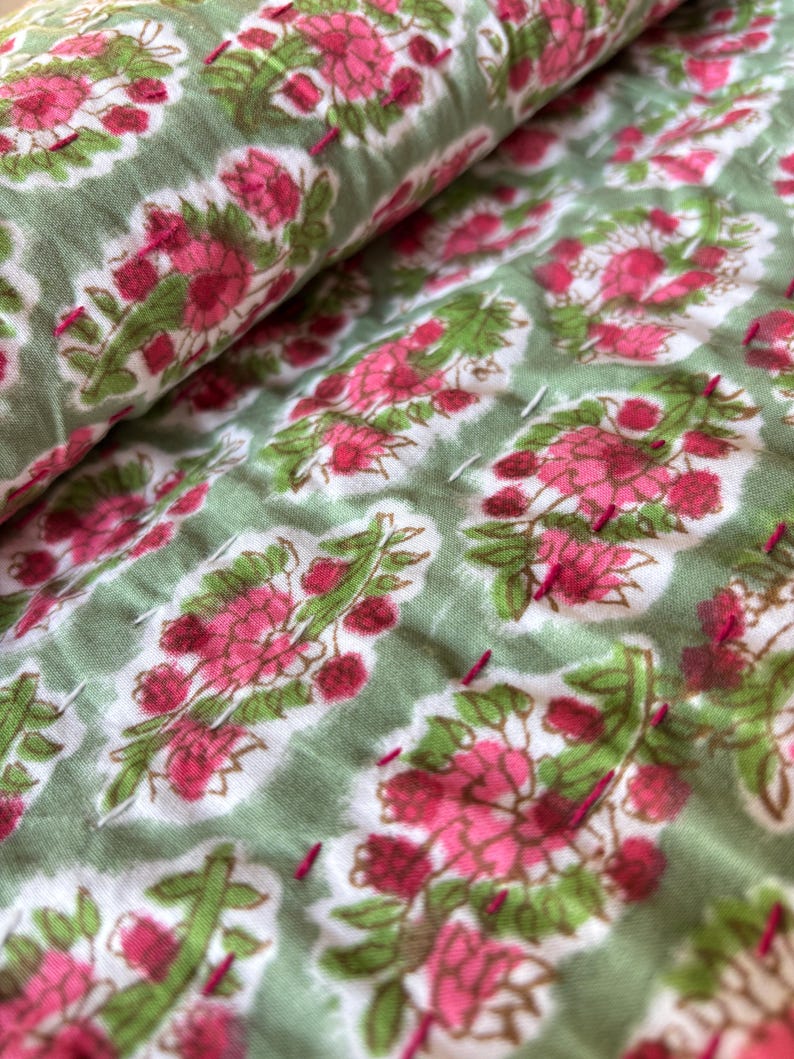May include: Close-up of a green fabric with a white background and a pink floral pattern. The fabric is quilted with pink thread.