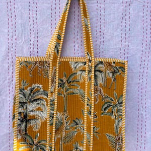 Puede incluir: Una bolsa de mano acolchada amarilla con ribete de rayas blancas y amarillas. La bolsa presenta un estampado de jungla con palmeras, jirafas y leopardos.