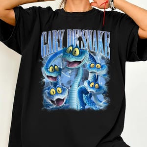Peut inclure: T-shirt noir avec un motif de serpents bleus de dessin anim&eacute; aux yeux jaunes. Le texte "GARY DESNAKE" est imprim&eacute; au-dessus des serpents dans une police bleue stylis&eacute;e. Le t-shirt a une coupe ample.