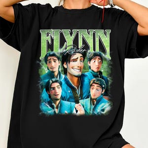 Puede incluir: Camiseta negra con un gráfico del personaje Flynn Rider de la película Enredados. El diseño incluye el nombre "Flynn Rider" en fuente verde, con múltiples imágenes del personaje. La camiseta es de Mira Medium Studio.