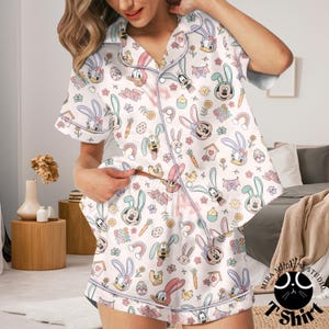 May include: White pajama set with an Easter-themed print featuring cartoon characters, bunnies, eggs, and rainbows. The short-sleeved top and shorts have light blue trim and the words "Easter" are visible on the shorts.