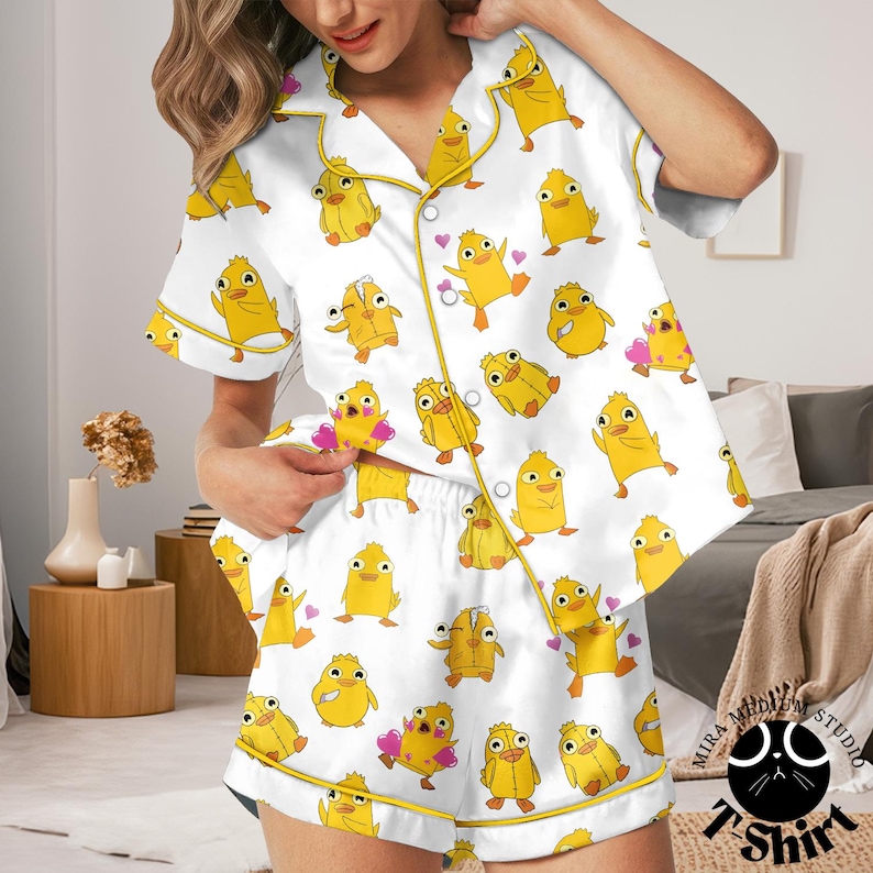 May include: White pajama set with a repeating pattern of yellow cartoon ducks and pink hearts. The short-sleeved top and shorts have yellow trim. The pajama set is made of a silky material.