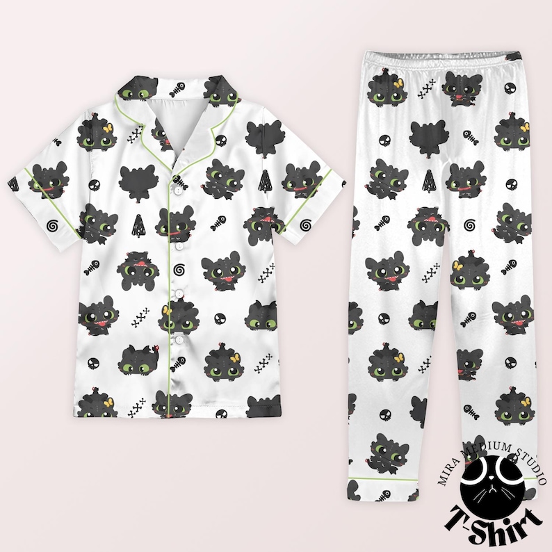 Puede incluir: Conjunto de pijama blanco con un estampado repetido de dragones de dibujos animados negros, espinas de pescado y otros peque&ntilde;os gr&aacute;ficos. La parte superior de manga corta tiene ribetes y botones verdes. El pantal&oacute;n de pijama tiene cintura el&aacute;stica.