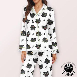 Puede incluir: Conjunto de pijama blanco con un patr&oacute;n repetido de dragones de dibujos animados negros, espinas de pescado y calaveras. El pijama tiene ribetes y botones verdes. La parte superior de manga larga y los pantalones son de material suave.