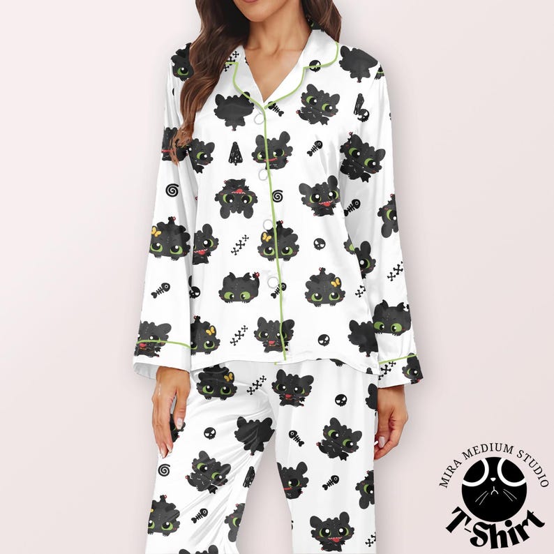 Puede incluir: Conjunto de pijama blanco con un patr&oacute;n repetido de dragones de dibujos animados negros, espinas de pescado y calaveras. El pijama tiene ribetes y botones verdes. La parte superior de manga larga y los pantalones son de material suave.