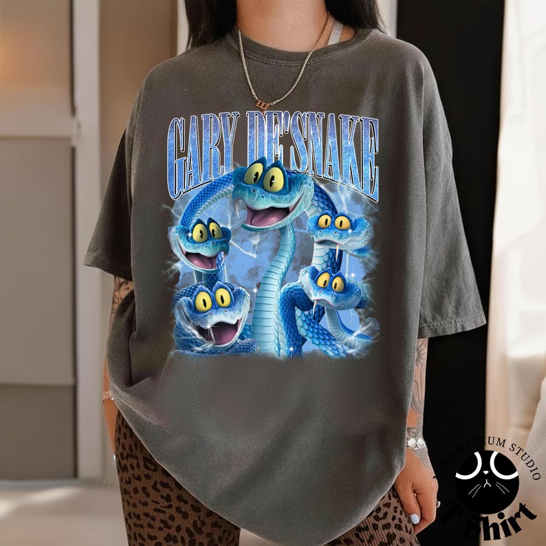 Peut inclure: T-shirt gris fonc&eacute; avec un motif de cinq serpents bleus aux yeux jaunes et le texte "GARY DE'SNAKE" dans une police bleue stylis&eacute;e. Les serpents ont la bouche ouverte et sont entour&eacute;s d'un motif bleu en forme d'&eacute;clair.