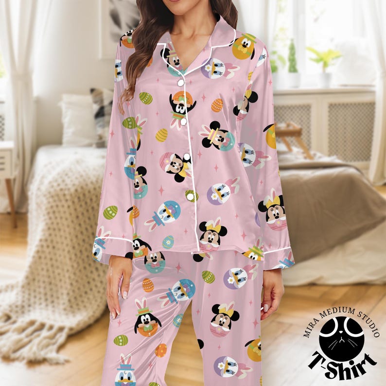 May include: Pink pajama set featuring a repeating pattern of cartoon characters in Easter-themed outfits. The long-sleeved top and pants have white trim and buttons. The pajama set is made of a soft, silky material.