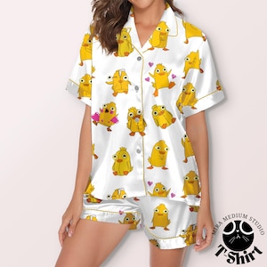 May include: White pajama set with a repeating pattern of yellow cartoon chicks with crowns and hearts. The short-sleeved top and shorts have gold trim. The pajama set is made of a silky material.