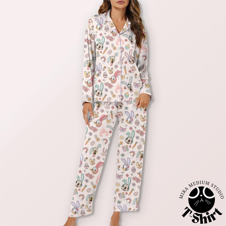 May include: White pajama set with an all-over Easter-themed print. The pattern includes cartoon bunnies, carrots, eggs, and flowers in pastel colors. The long-sleeved top has a notched collar and button-down front. The pants are straight-legged.