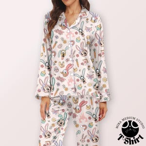 May include: White pajama set with a repeating pattern of cartoon bunnies, eggs, carrots, and castles. The long-sleeved top has a button-down front and a notched collar. The pajama bottoms have an elastic waistband. The set is made of a silky material.