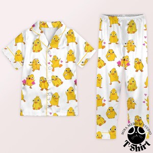 May include: White pajama set with a repeating pattern of yellow cartoon ducks and pink hearts. The short-sleeved top has yellow trim and buttons. The matching pants complete the set, perfect for a comfortable night's sleep.
