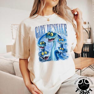Peut inclure: T-shirt cr&egrave;me avec une illustration de dessin anim&eacute; de serpents bleus aux yeux jaunes. Le texte "GARY DE'SNAKE" est arqu&eacute; au-dessus des serpents. Le t-shirt a des manches courtes et un col rond.
