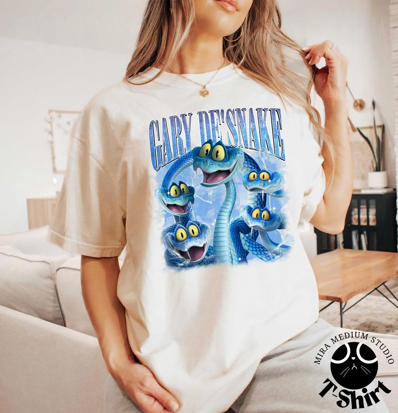 Peut inclure: T-shirt cr&egrave;me avec une illustration de dessin anim&eacute; de serpents bleus aux yeux jaunes. Le texte "GARY DE'SNAKE" est arqu&eacute; au-dessus des serpents. Le t-shirt a des manches courtes et un col rond.