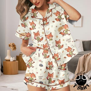 May include: Cream-colored pajama set featuring a repeating pattern of cartoon foxes in various poses, with text bubbles and phrases like "Smart Fox" and "Wild Times." The set includes a button-down shirt and shorts, both trimmed in brown.