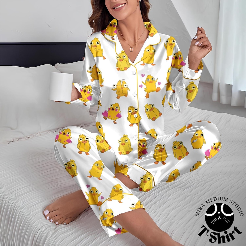 May include: White pajama set with a repeating pattern of yellow cartoon chicks. The long-sleeved top and pants have yellow trim. The pajama set is made of a silky material. The image also includes a white mug and a logo that says "Mira Medium Studio T-Shirt".
