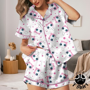 Könnte beinhalten: Weißes Satin-Pyjama-Set mit einem sich wiederholenden Muster aus rosa Cartoonfiguren und Sternen. Das Set beinhaltet ein kurzärmeliges Hemd mit Knöpfen und passende Shorts, beide mit rosa Paspeln versehen.