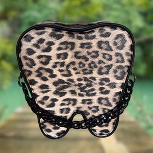 May include: A novelty handbag shaped like a tooth, featuring a leopard print pattern in shades of tan and black. The bag has a black zipper and a black chain strap. The design is unique and eye-catching.