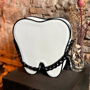 May include: White tooth-shaped handbag with a black chain strap. The bag has a black trim around the edges.