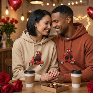 Könnte beinhalten: Ein Paar trägt passende Kapuzenpullover, einer beige, einer rostrot, mit einem roten Roller-Motiv. Sie sitzen an einem Tisch mit Kaffeetassen, Pralinen und roten Rosen. Herzförmige Ballons im Hintergrund.