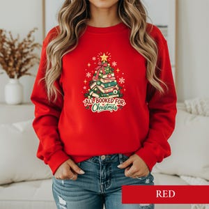 Op de afbeelding: Een rood sweatshirt met lange mouwen en een afbeelding van een kerstboom gemaakt van gestapelde boeken. Het ontwerp bevat een gele ster als piek, een rode strik en de tekst "ALL BOOKED FOR CHRISTMAS" in rode en groene letters.