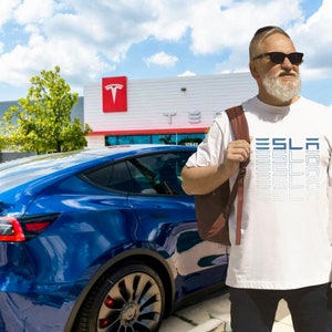 Könnte beinhalten: Ein weißes T-Shirt mit dem Wort "TESLA" in einem gestapelten, abgestuften Design. Die Person trägt eine Sonnenbrille und einen braunen Rucksack. Ein blaues Tesla-Auto parkt vor einem Tesla-Autohaus.