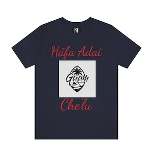 Op de afbeelding: Donkerblauw T-shirt met rode tekst die "Hafa Adai Guam Che'lu" zegt. Het shirt heeft een wit vierkant met een zwart-witte afbeelding van de vlag van Guam.