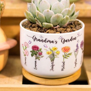 May include: White ceramic planter with a succulent plant and the text "Grandma's Garden". The planter has a wooden base and features five different flowers with names written below each flower: James, Emma, Jennifer, Michael, and Sophia.