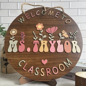 May include: Welcome to Ms. Taylor's Classroom sign with a brown wooden background and colorful letters. The sign features a heart and flowers in pots.