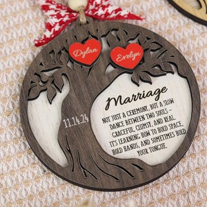 May include: A wooden, circular ornament with a tree design, featuring the names "Dylan" and "Evelyn" on red hearts. The ornament includes the date "11.14.24" and the word "Marriage" with a quote. It is adorned with a red and white bow.