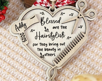 May include: A heart-shaped wooden Christmas ornament with the text "Blessed are the Hairstylists" and a comb, scissors, and a hairdryer design. The ornament has a red and white bow and the name "Addy" and the year "2025".