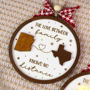 May include: A round wooden Christmas ornament with a white center and a brown wooden border. The ornament features the text "THE LOVE BETWEEN FAMILY KNOWS NO DISTANCE" and two state outlines connected by a dotted line.