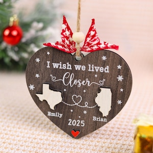 May include: A heart-shaped wooden Christmas ornament with the text "I wish we lived Closer" and the names "Emily" and "Brian". The ornament features the states of Texas and Illinois, with a red heart at the bottom. A red and white ribbon is at the top.